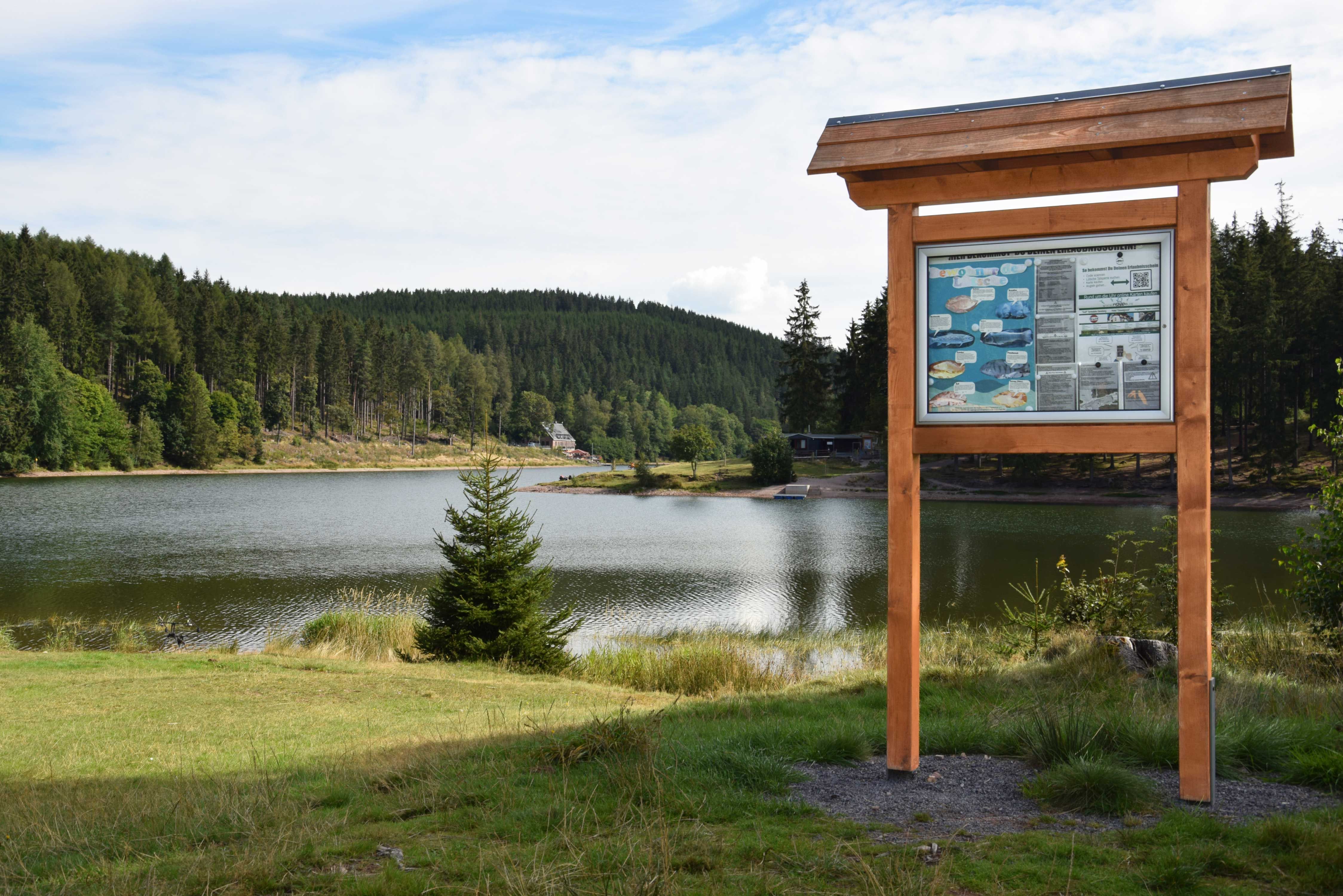 Ein informatives Holzschild steht im Uferbereich der Talsperre Lütsche, dahinter liegt der Badesee.